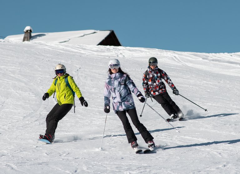 Un groupe de 3 amis déscend une piste avec un chalet dont le toît est enneigé en fond